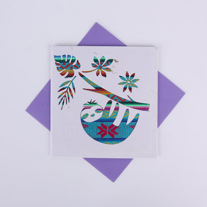 Colourful patterned cutout on a white card showing a sloth hanging from a tree with a purple envelope underneath